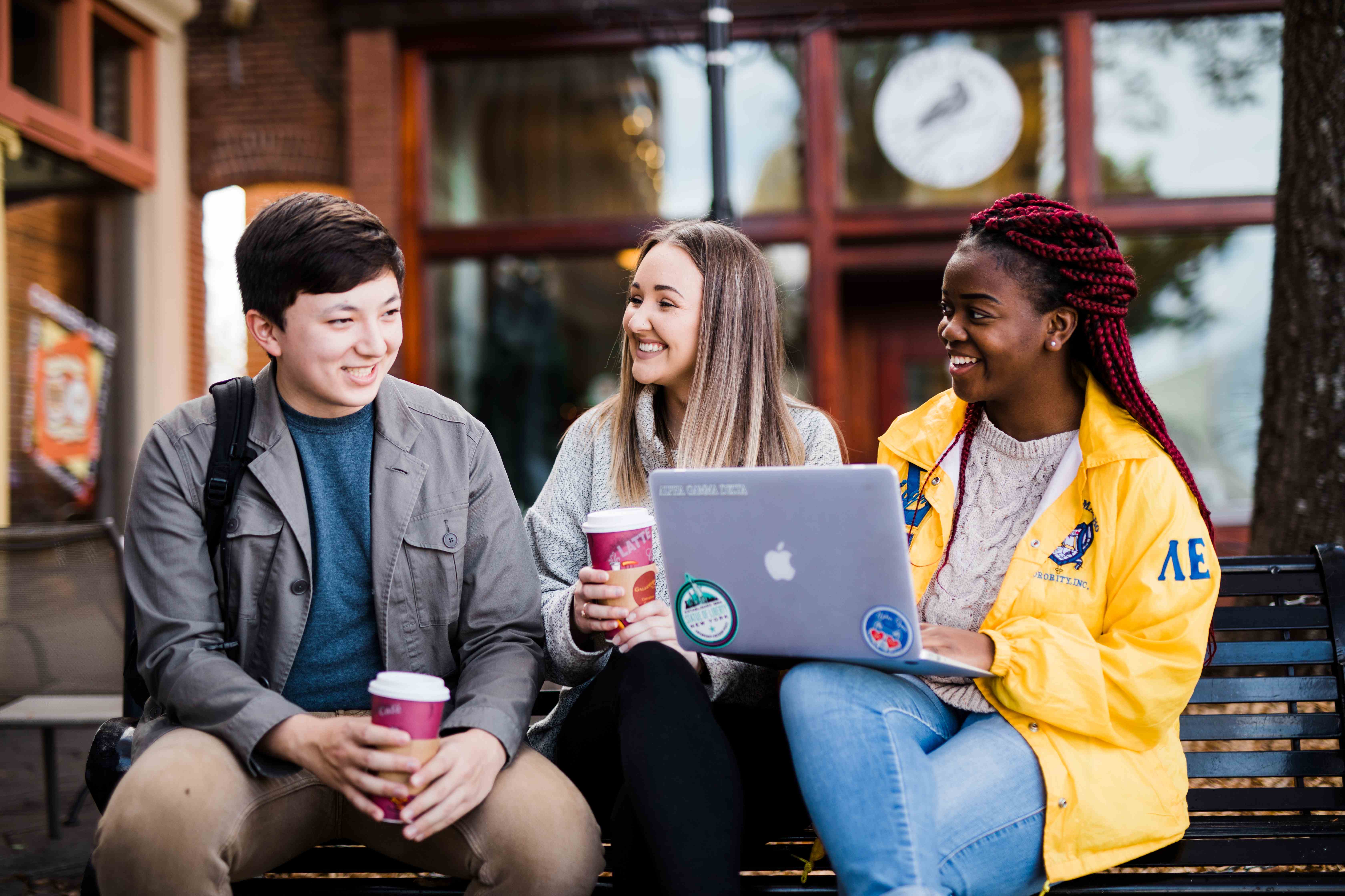 3 students sitting outside while talking and drinking coffee