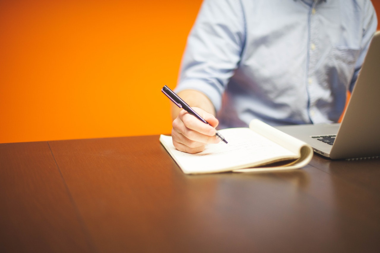 Person Writing on Note Beside Gray Laptop Computer