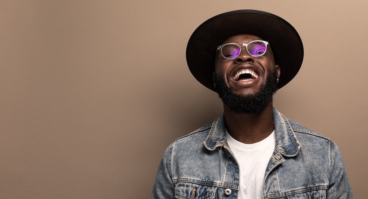 Black man in jean jacket, hat, and shades smiling at the camera