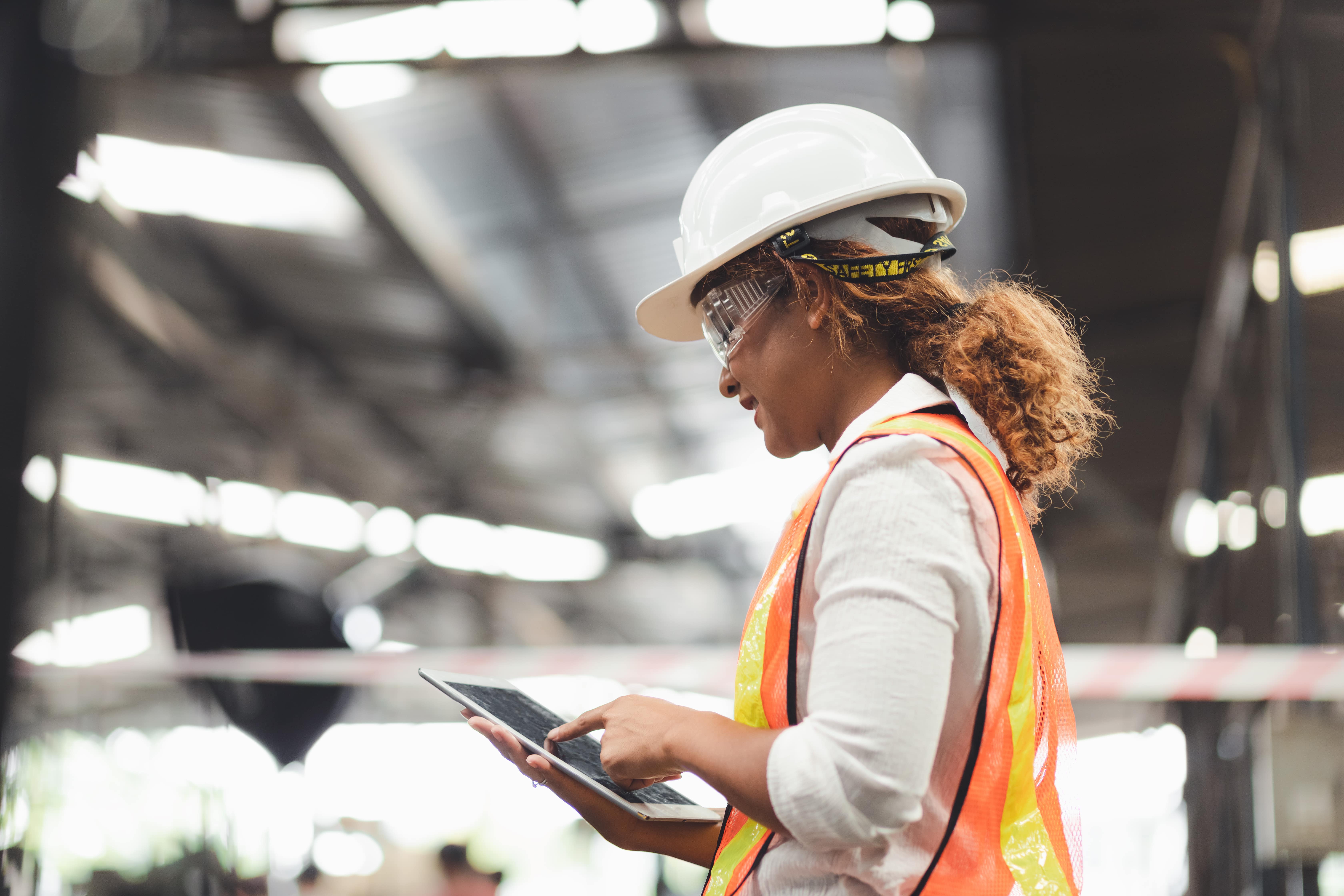 Female wearing construction vest and hard hat, looking at a tablet and standing in a warehouse building.