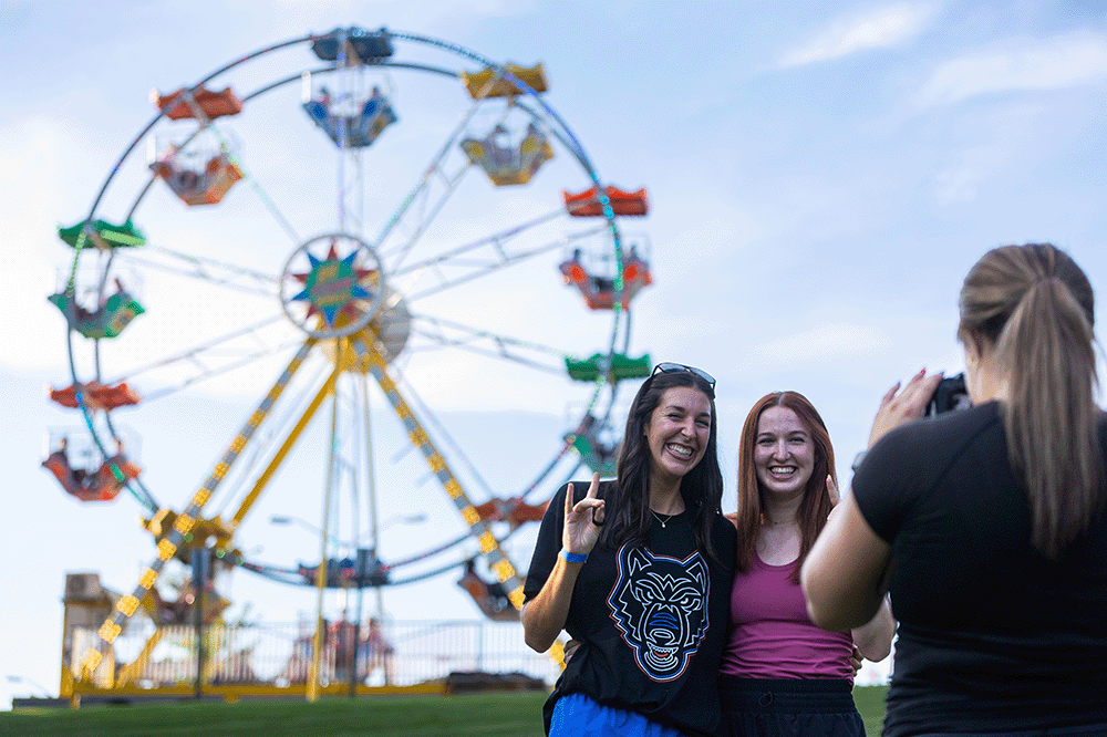 Two people posing for a photo in front of a Ferris wheel at a fair while another person takes the picture.