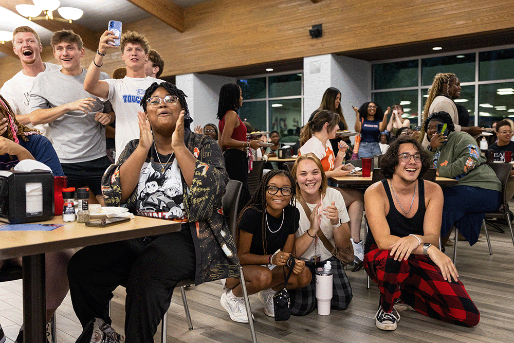 Group of people gathered in a dining hall, sitting and standing around tables while watching and reacting to an event