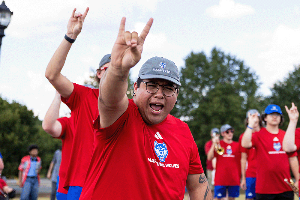 Person in a red shirt making a hand gesture toward the camera while a group wearing matching red shirts stands outdoors behind them, some holding instruments