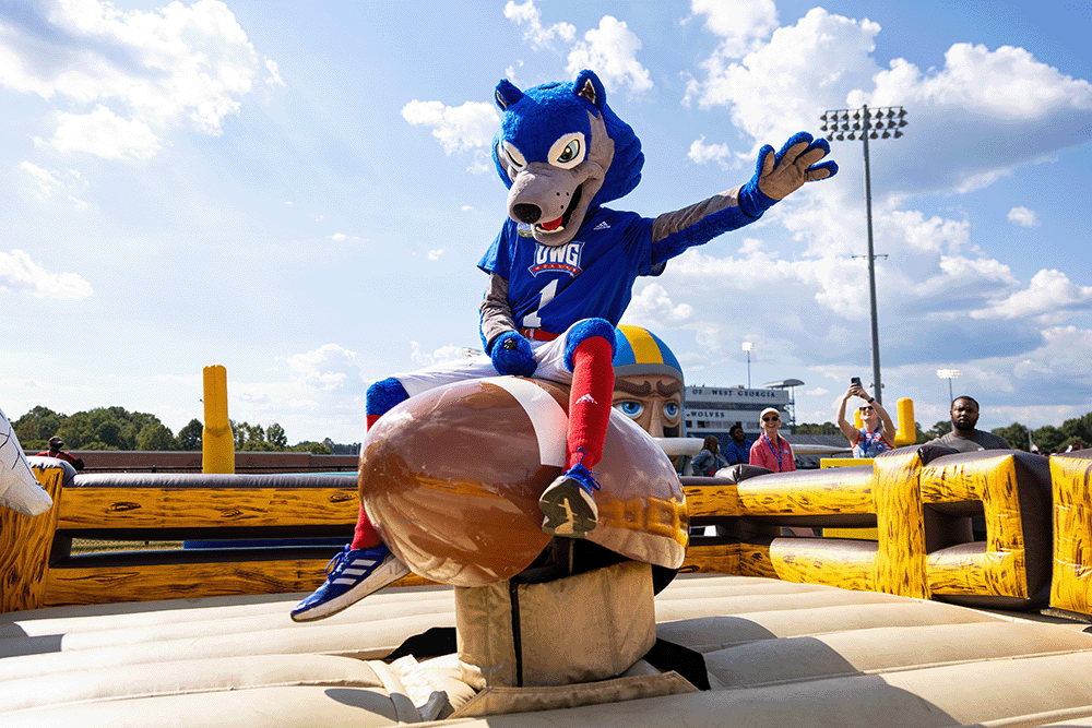 University mascot Wolfie in a blue uniform sitting on a mechanical bull ride at an outdoor event with people and stadium lights in the background