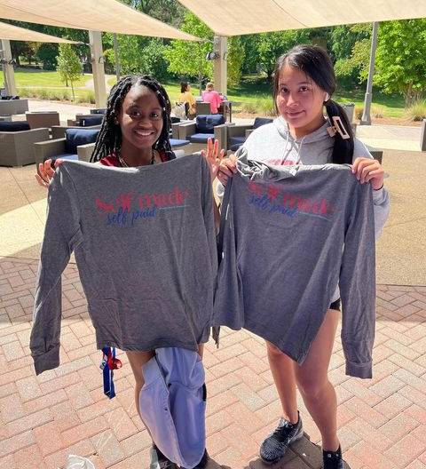 Two students standing outside of the Campus Center holding up shirts saying "Self Made, Self Paid".