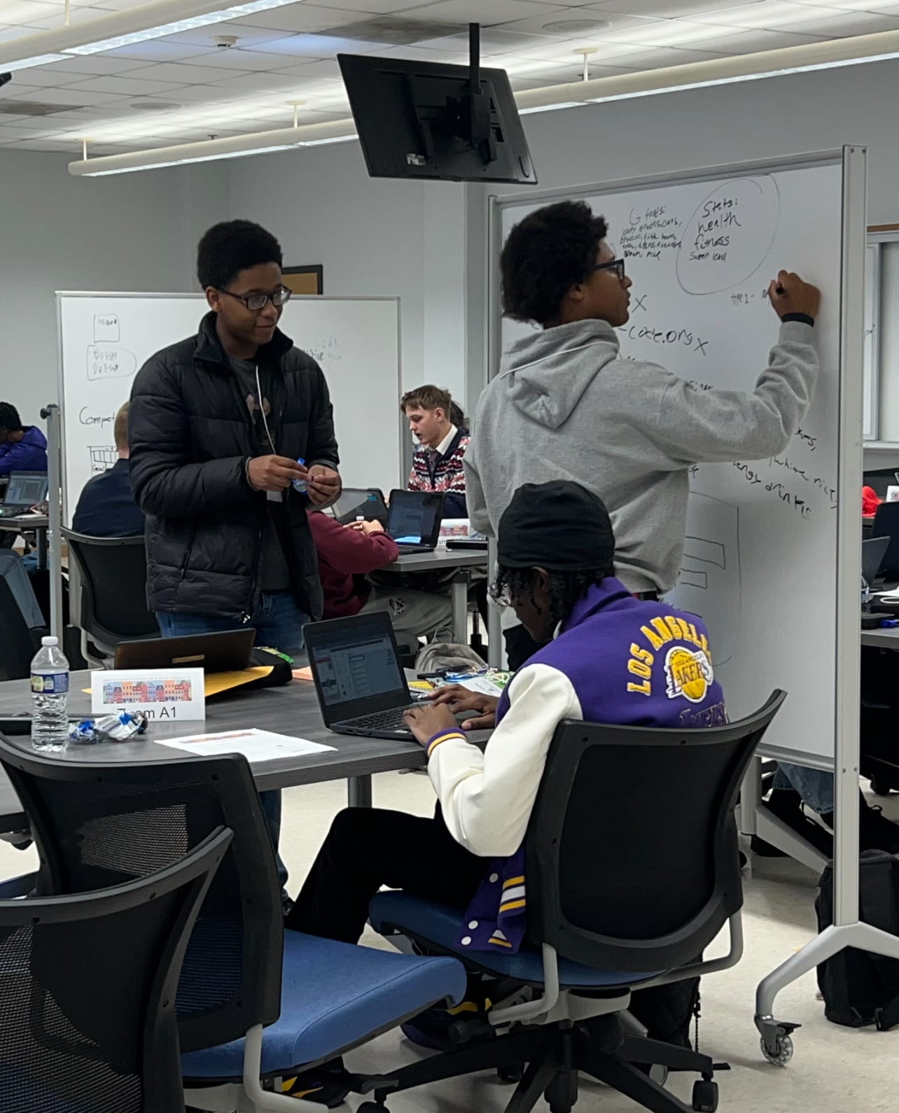 students writing on white board with team mates working on laptop