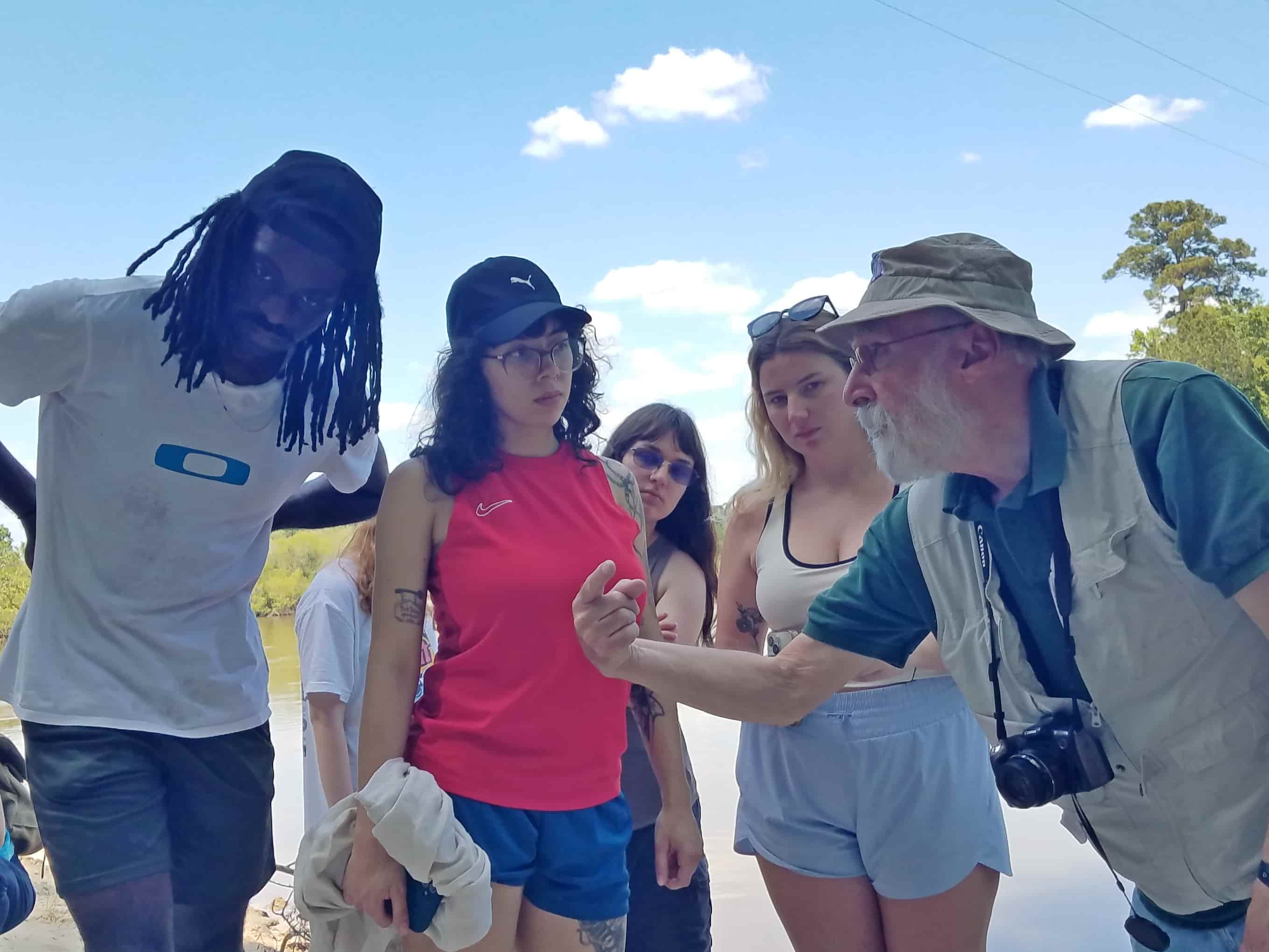 Dr. Chowns explaining a field study concept to a small group of students gathered outdoors near a body of water during a hands-on learning activity.