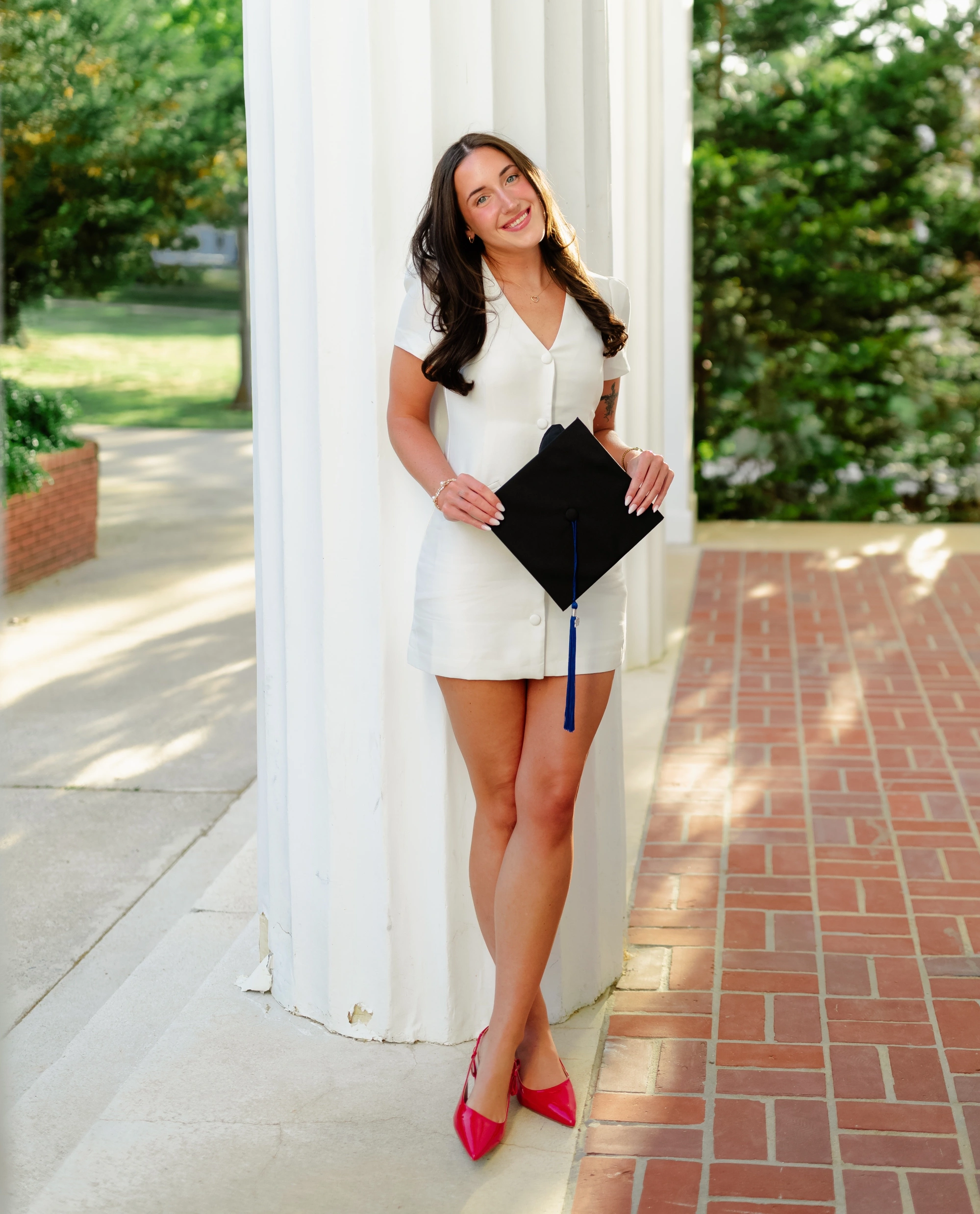 Rylie wearing a white dress and red shoes, holding her graduation cap and standing in front of a white column at the entrance to the Old Auditorium on campus.