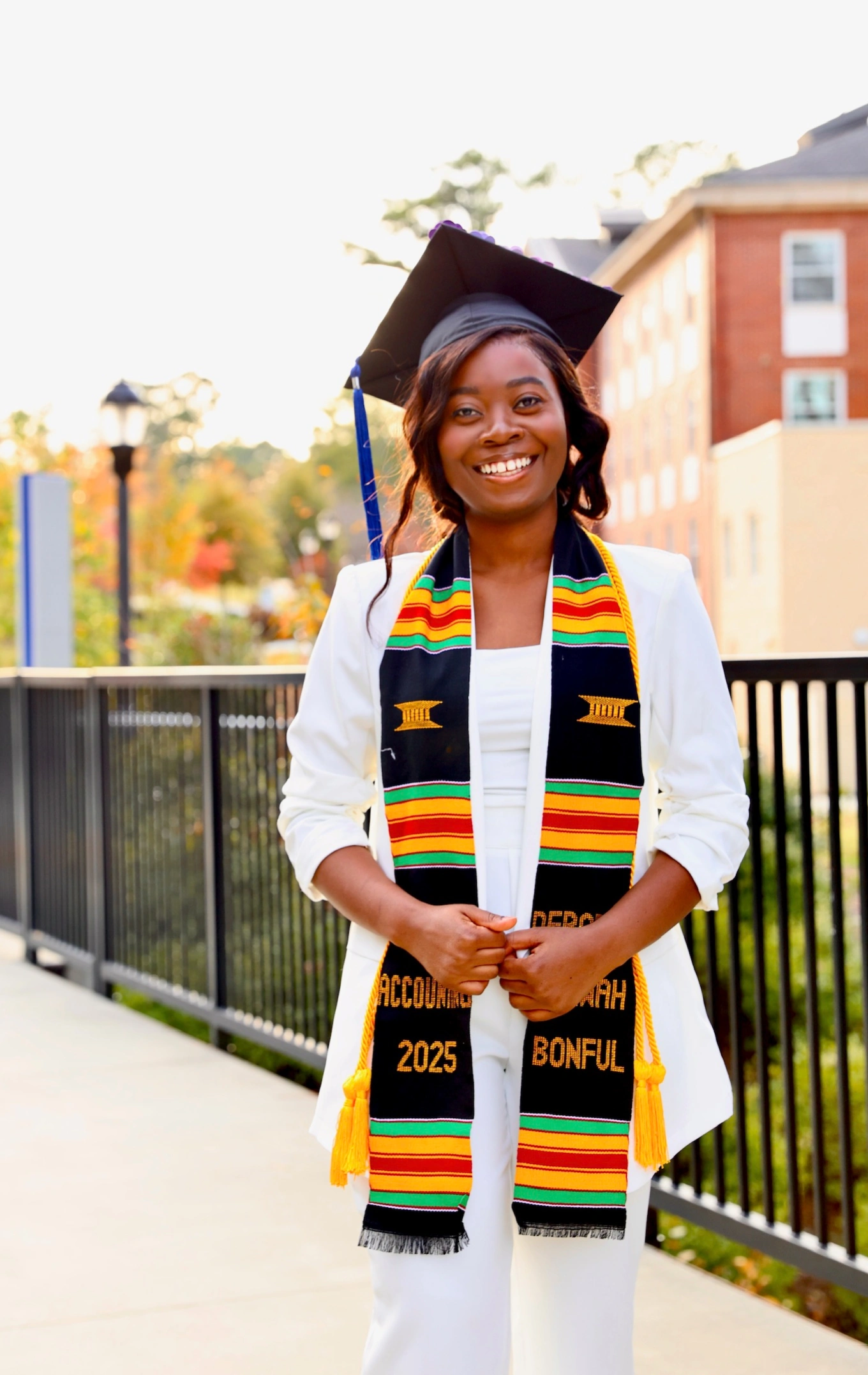 Deborah standing in front of the Richards College of Business in a white suit, cap, and wearing her stole.