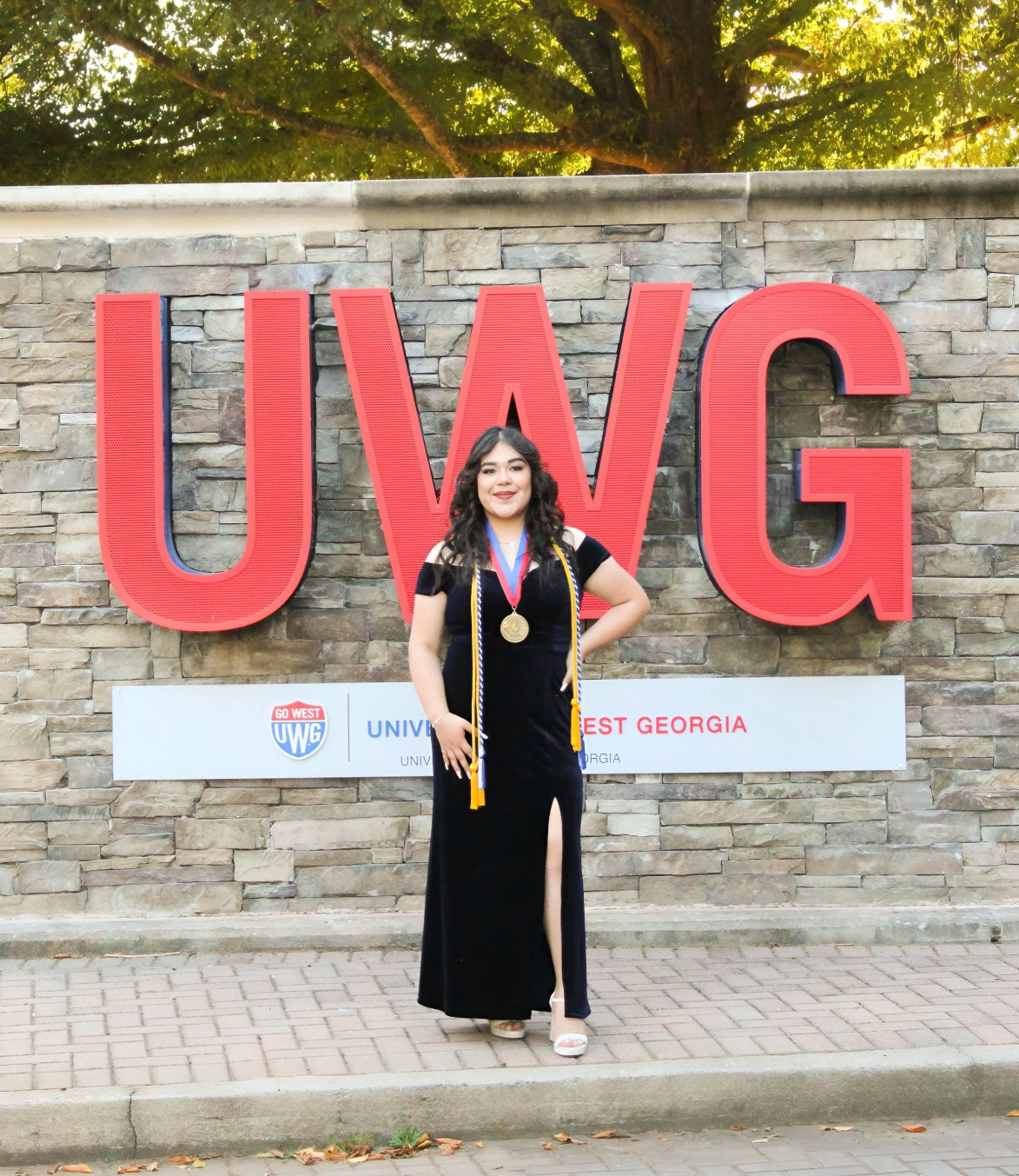 Lina standing in front of UWG's entrance sign wearing a black dress and her cords.