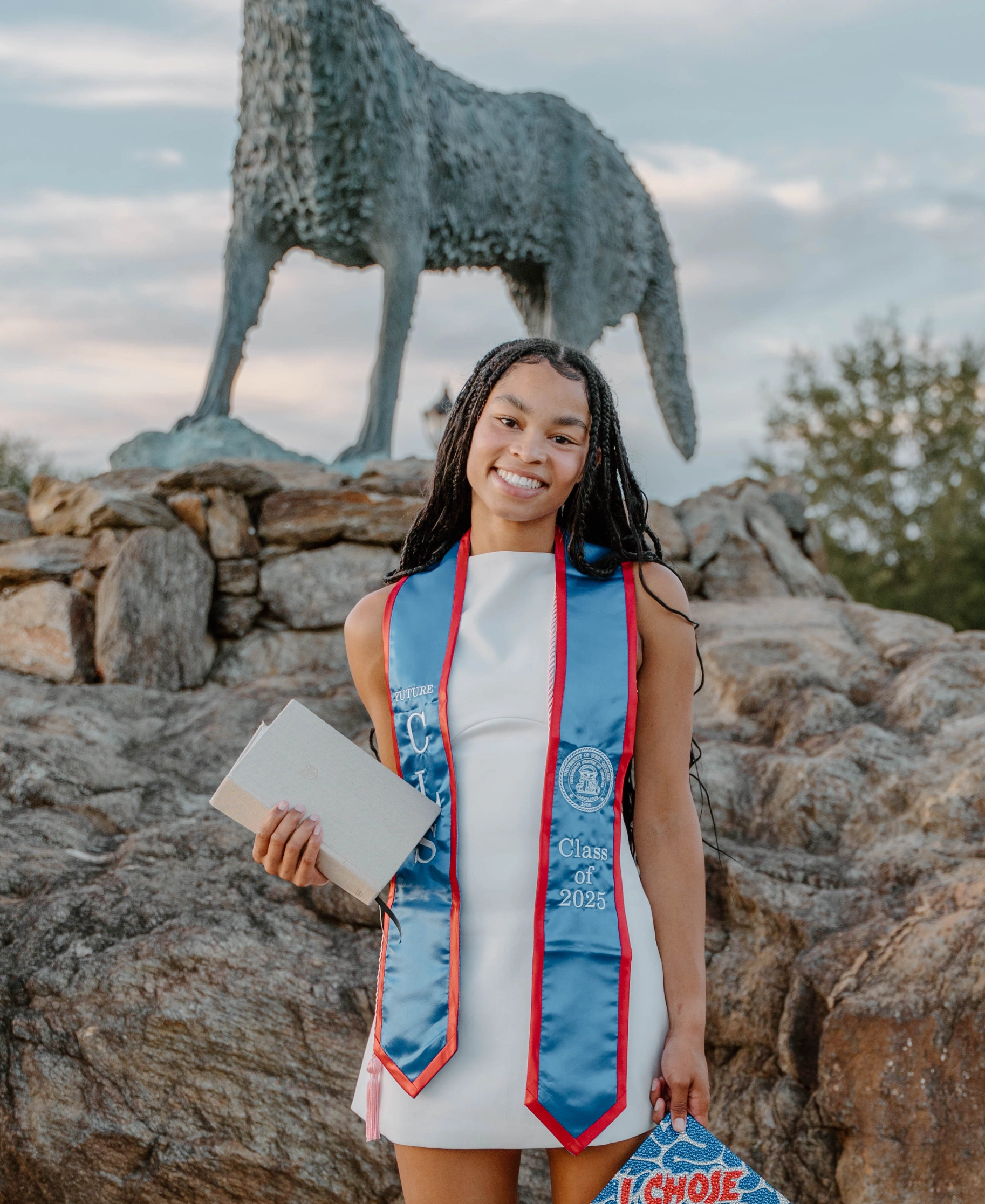 Kaitlyn wearing a white dress and holding a notebook and her cap, which she customized with red and blue gems, posing in front of the Wolf Plaza statue.