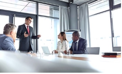 Man in suite standing at the head of a conference table talking to his team.