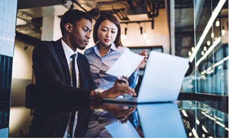 Two people in business attire sitting at a computer and reviewing a document.