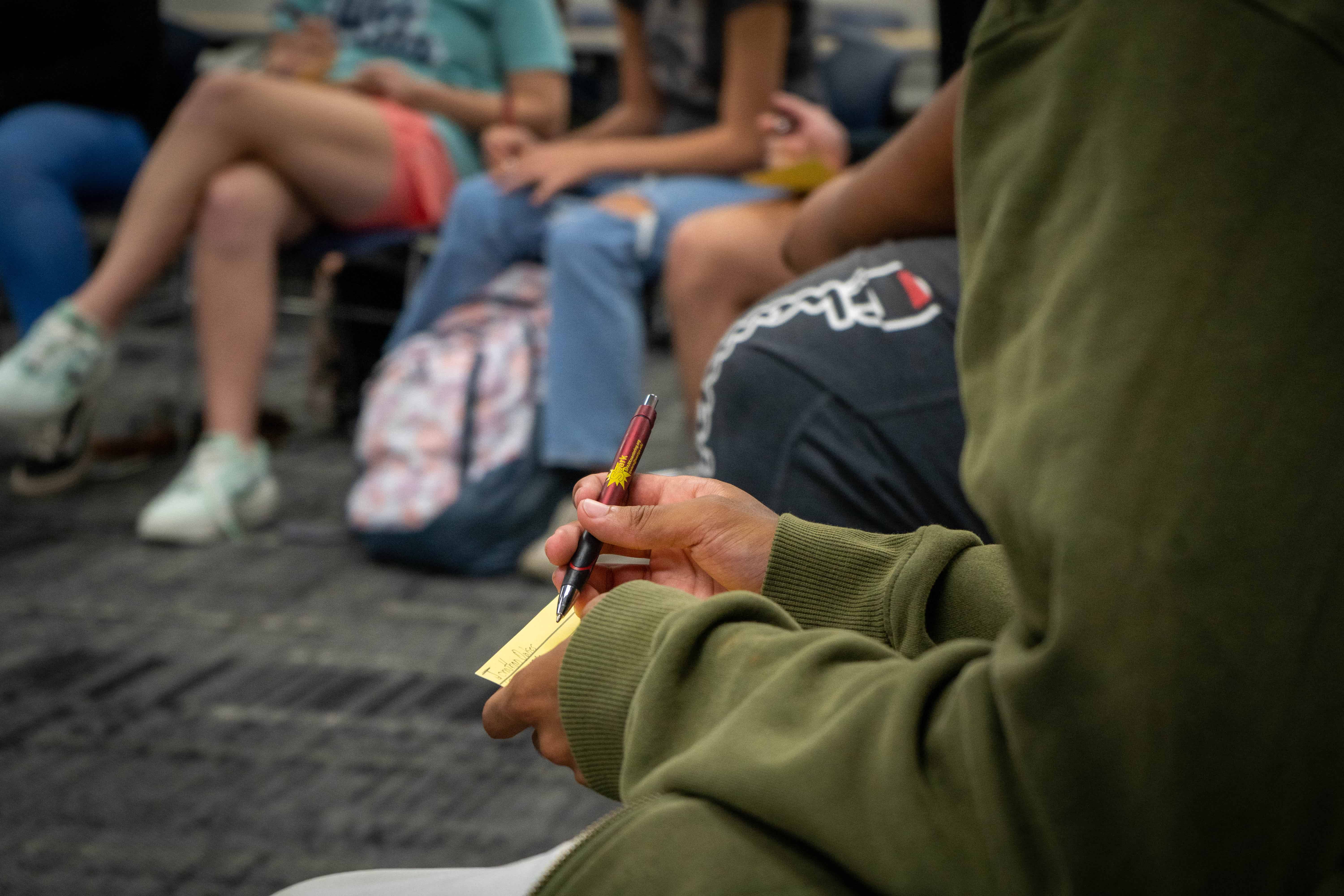 Students sitting in a circle with camera focused on one student writing a note.