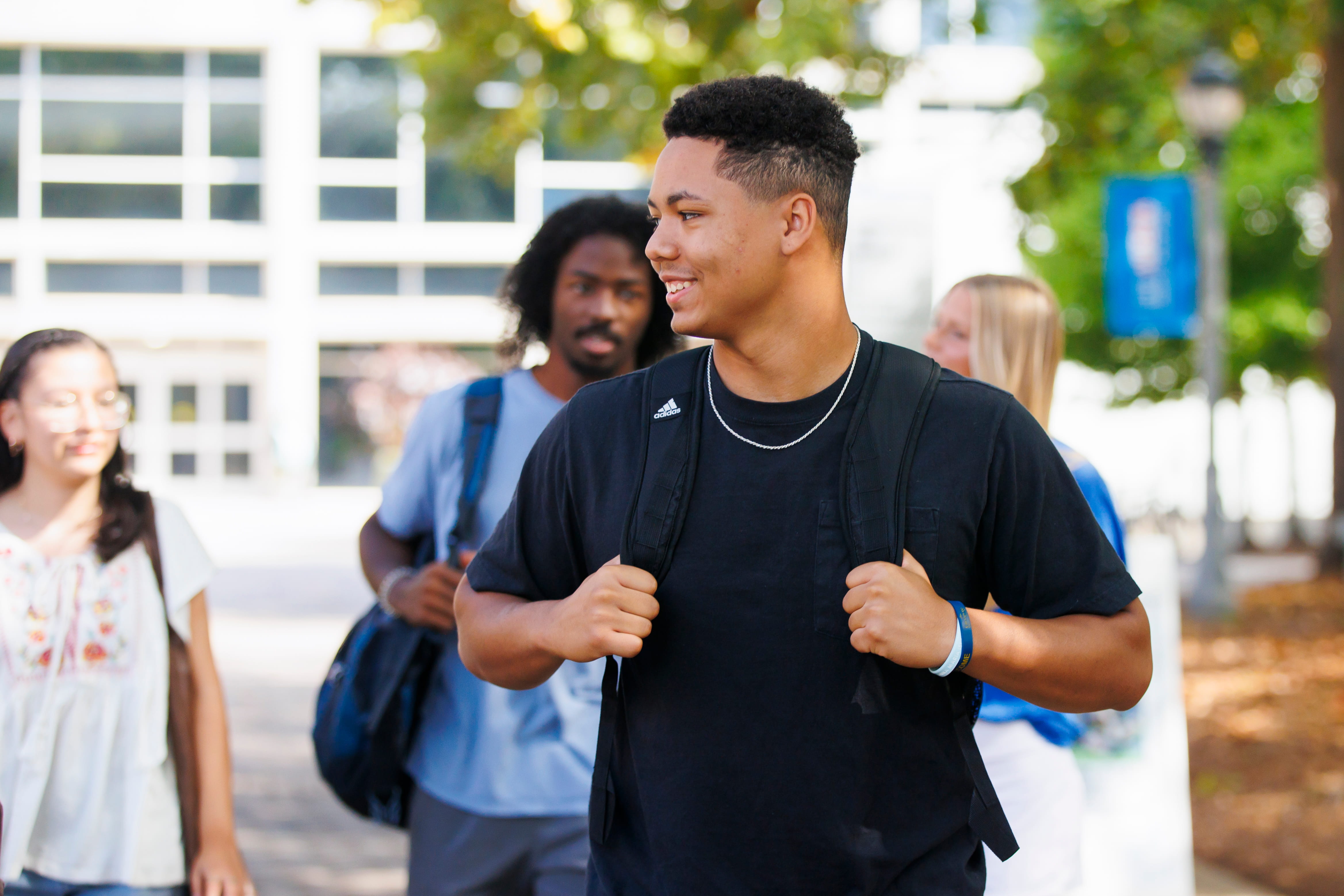 Student walking and smiling while holding his backpack straps.