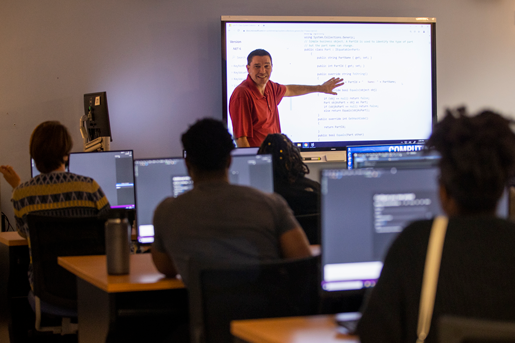 Photograph of an instructor gesturing towards a project in front of a class of students.