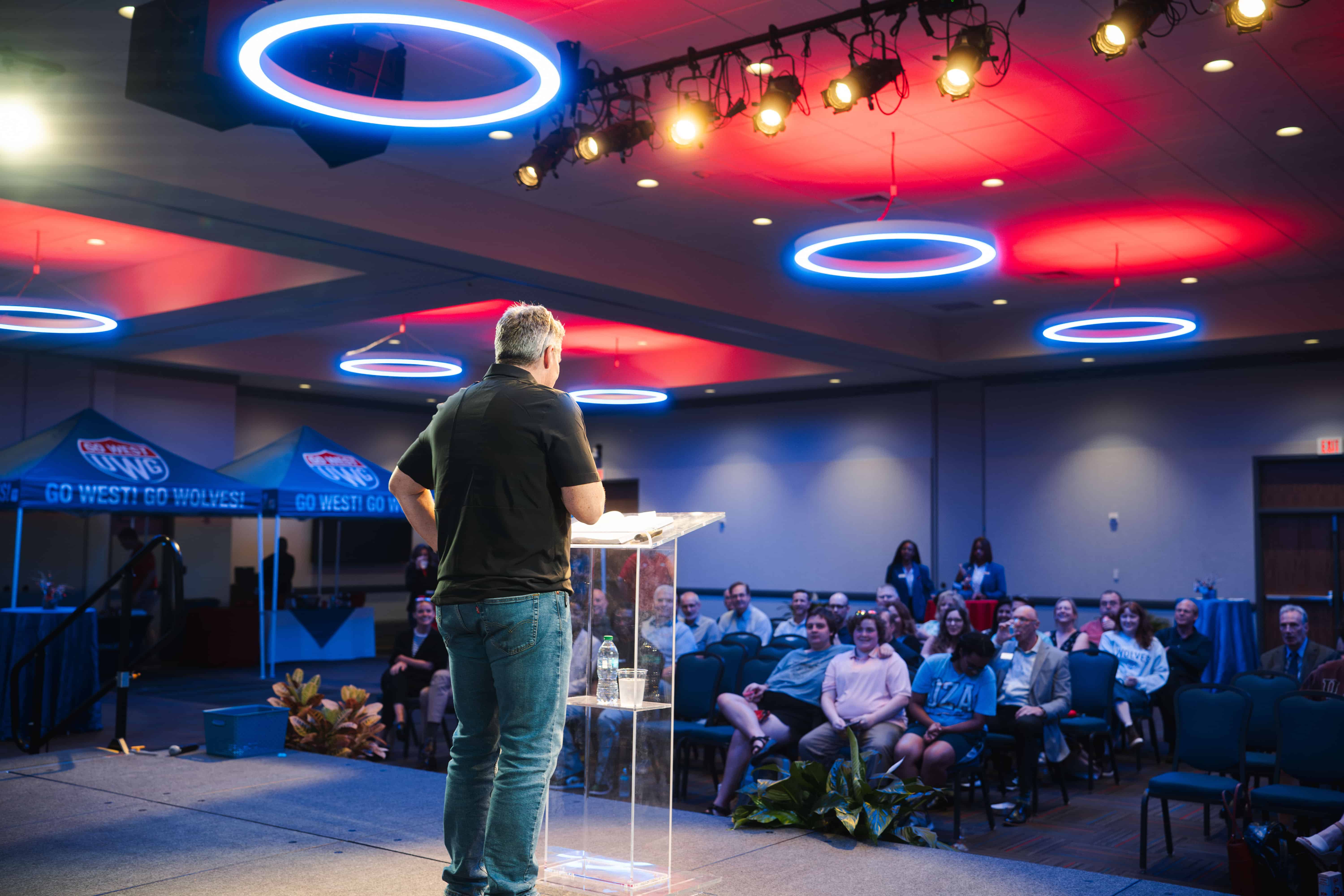 Presenter speaking in front of a group of listeners in the Campus Center Ballroom with red and blue lights overhead.