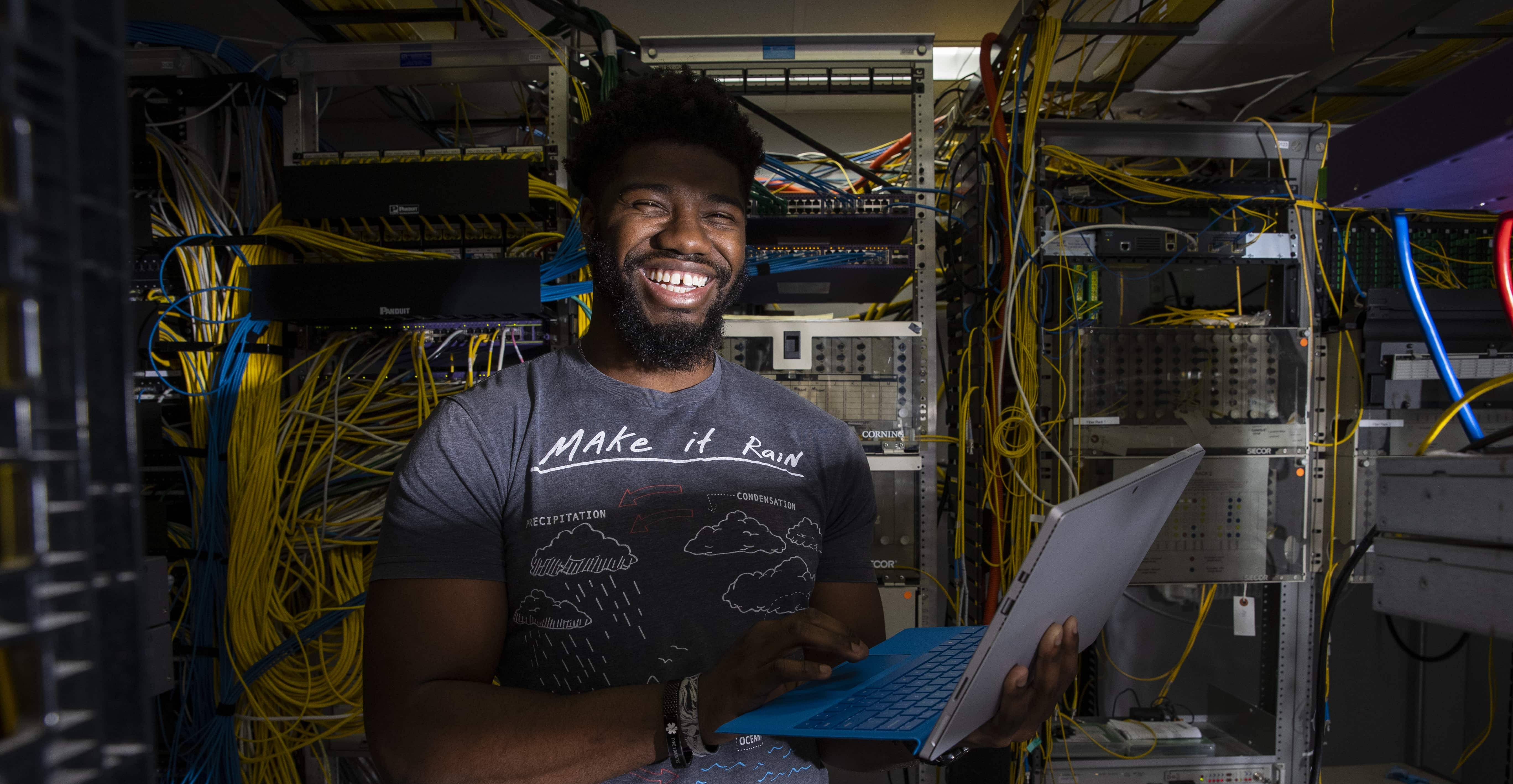 Student with laptop smiling in server room with cables all around.