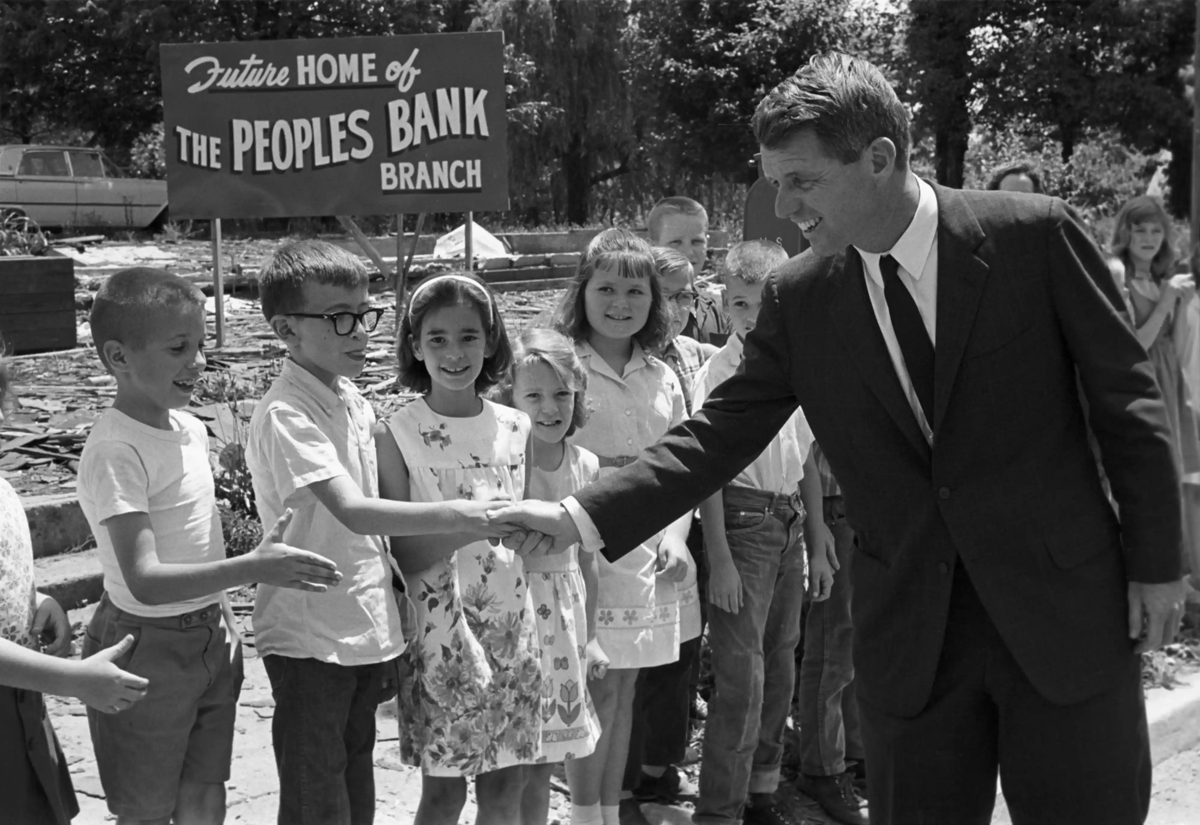 Historic black and white photo of President Kennedy shaking hands with a group of children.