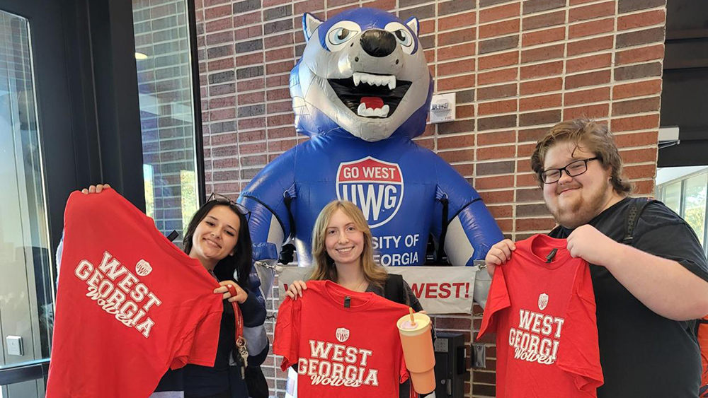 Three Students holding up red "West Georgia Wolves" T-shirts smiling in front of an inflatable Wolfie on Newnan Campus