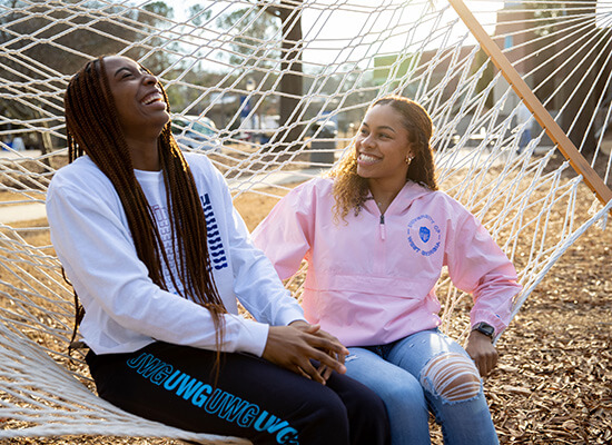 Two laughing UWG students sitting in a hammock outside on campus
