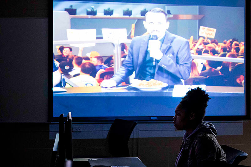 A student sits in front of a projector screen showing a gaming commentator