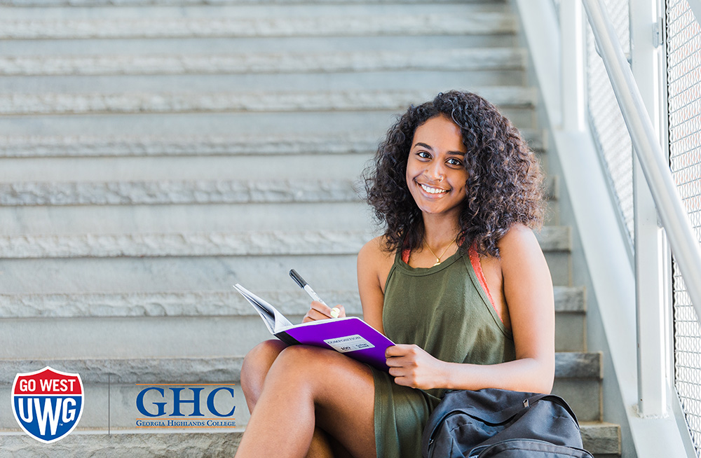 Photo of a student writing in a notebook with the UWG and GHC logos in the left bottom corner 