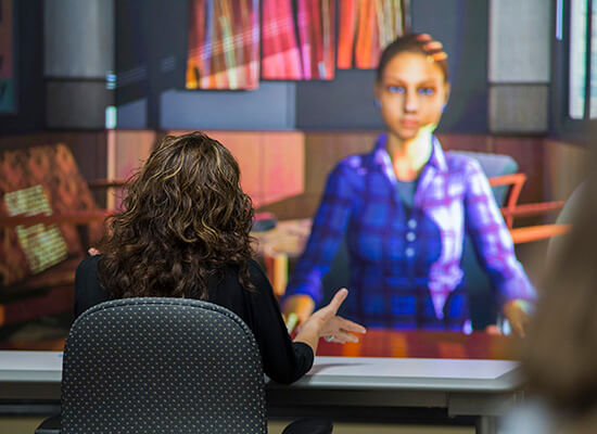 Person sitting at a table facing a screen with an avatar displayed