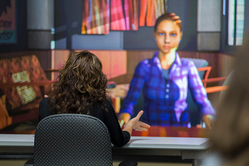 Person sitting at a table facing a screen with an avatar displayed on it