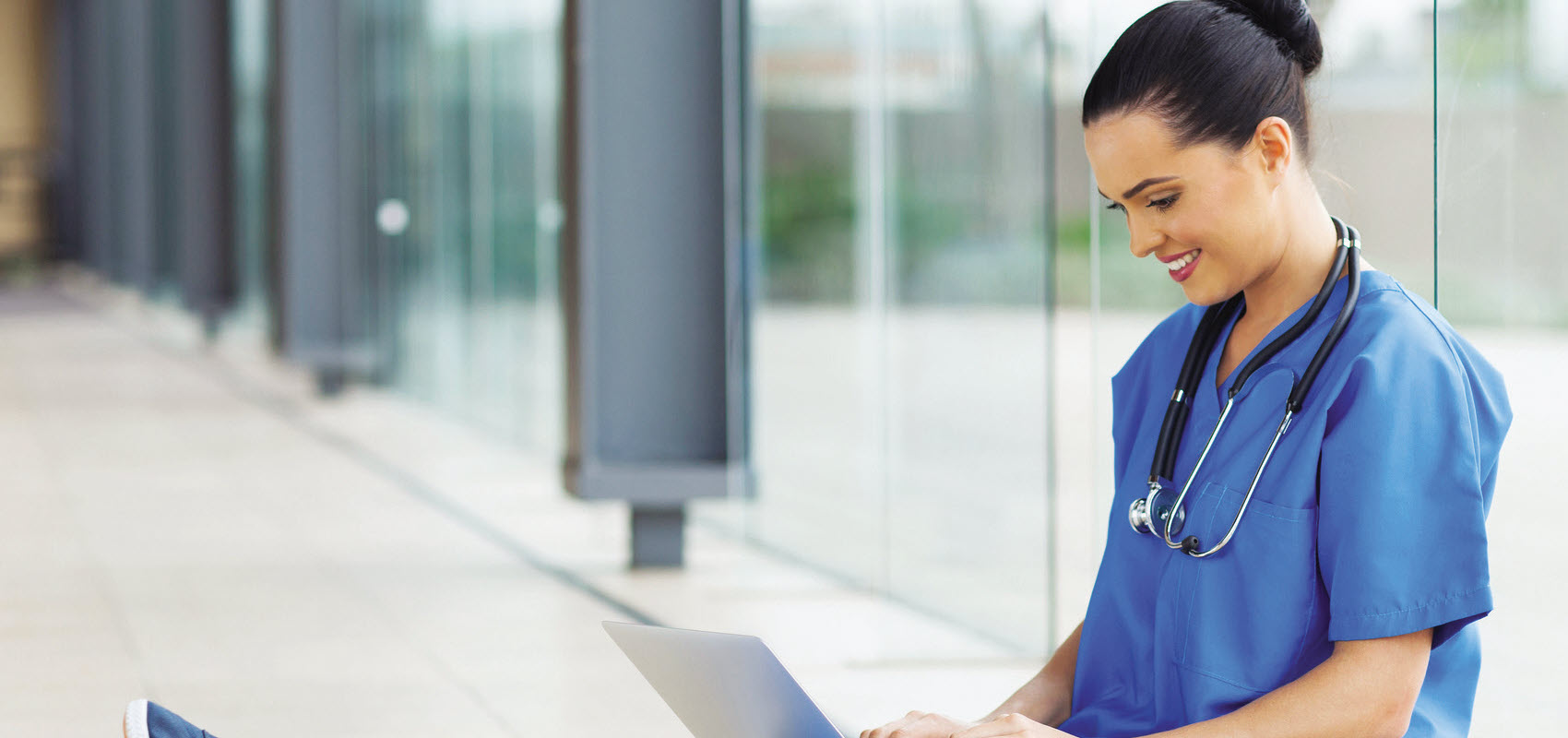 Nursing student in blue scrubs sitting against a window wall on her computer.