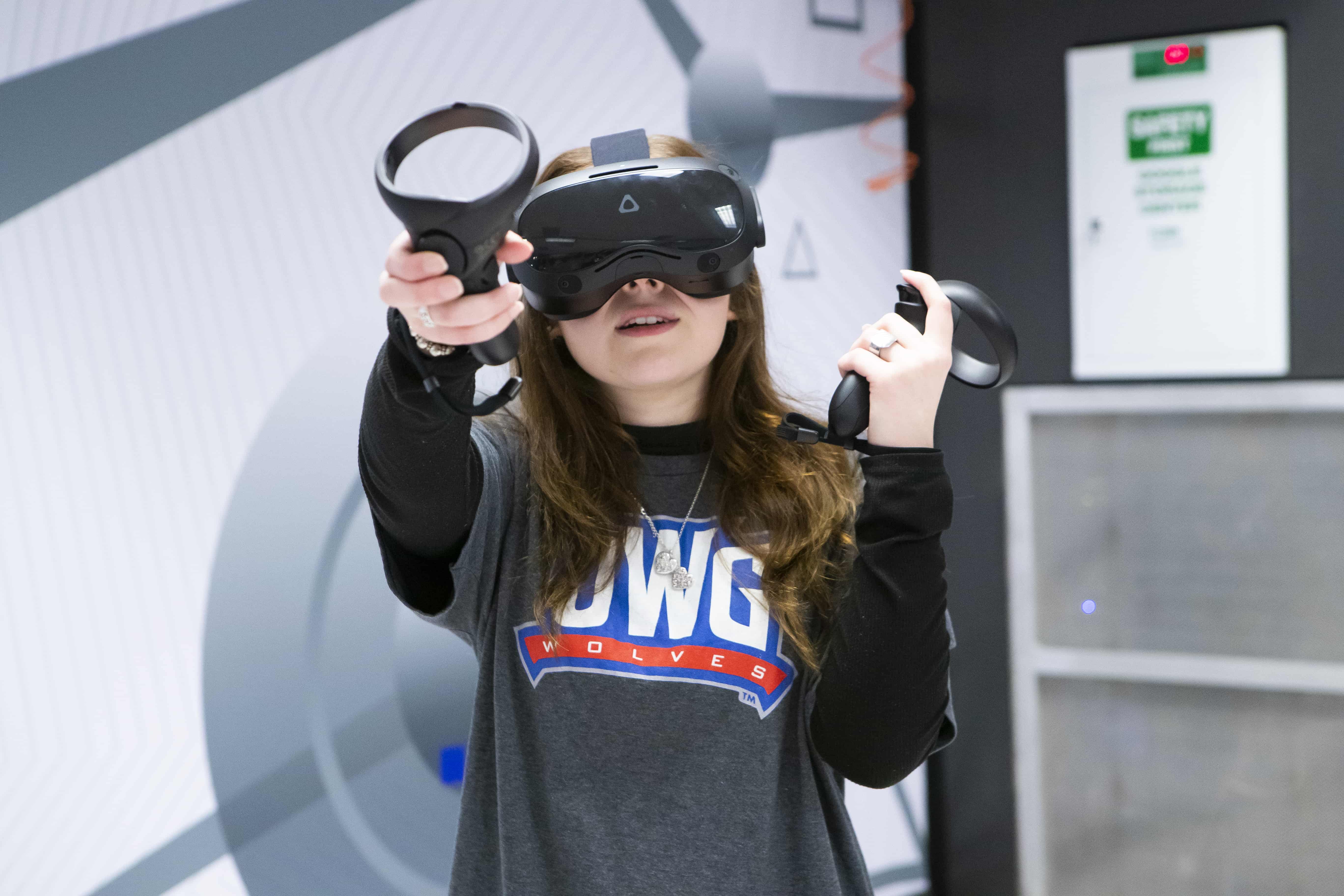 Female student wearing virtual reality gear, using the controller to point upwards while inside the VR interactive space.
