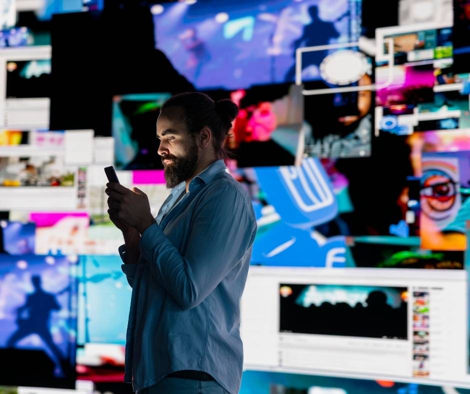 Man illuminated by a smartphone screen while surrounded by a wall of vibrant digital media content