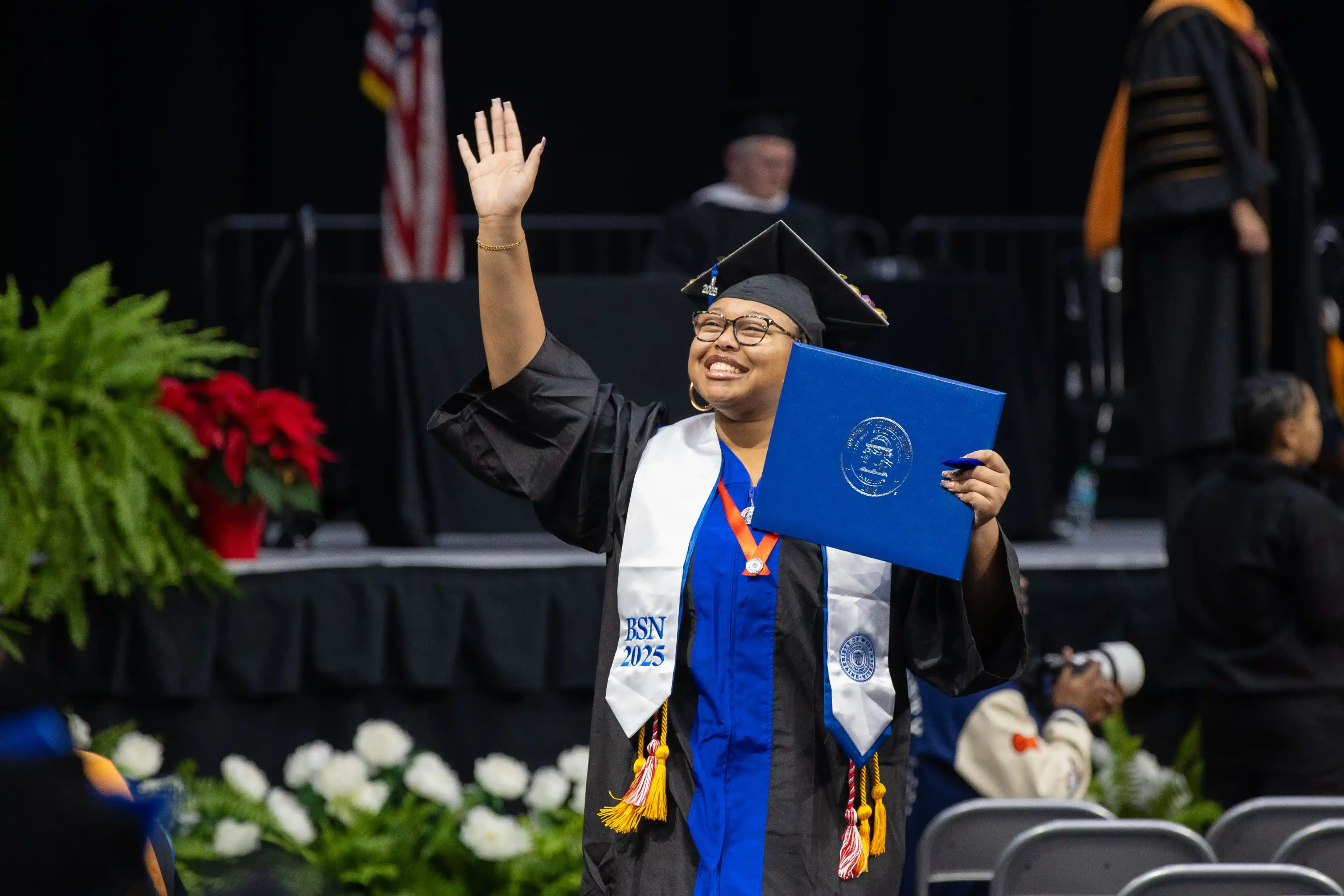 Student holding up deploma and waving to crowd smiling while walking at commencement.