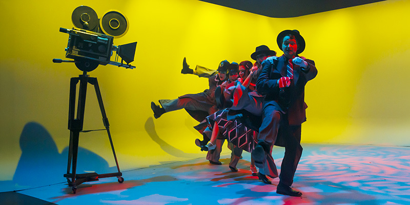 People in vintage-style clothing performing a synchronized high-kick dance in a brightly lit yellow studio, with an old film camera on a tripod nearby.