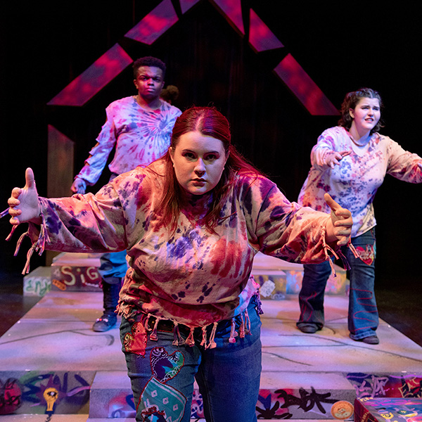 Three performers in tie-dye costumes reach forward in a dramatic pose on a colorful stage with patterned steps and geometric lighting.