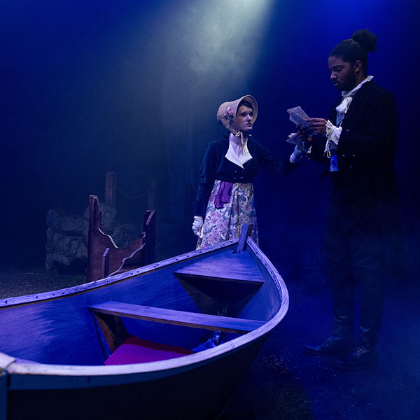 Two performers in period costumes stand beside a wooden rowboat on a dim, blue-lit stage while one reads from papers and the other looks on.