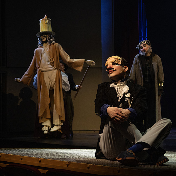 Performer in period costume sits on a stage foreground wearing a stylized mask, with other costumed performers and a puppet-like figure.