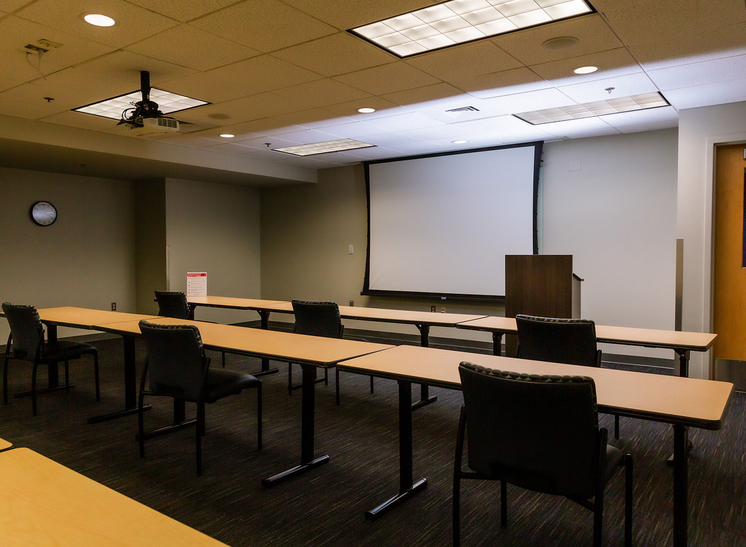 Meeting Room 302 facing the front of the room showcasing long tables with spaced seating, a podium, and a projector screen