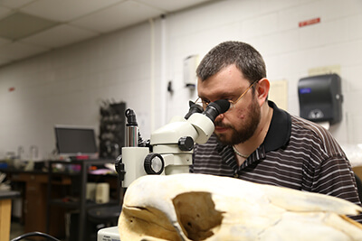 Christian Bryant sits and gazes through the dual eyepieces of a microscope in a labroom.