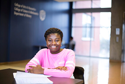 Jesica Sarfo is seated with her notebook, arms crossed, in a large lobby at UWG.