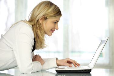 Woman in white jacket leaning on her desk looking at her laptop.