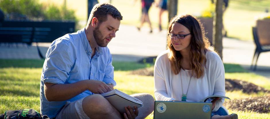 Students sitting outside with laptops and textbooks.